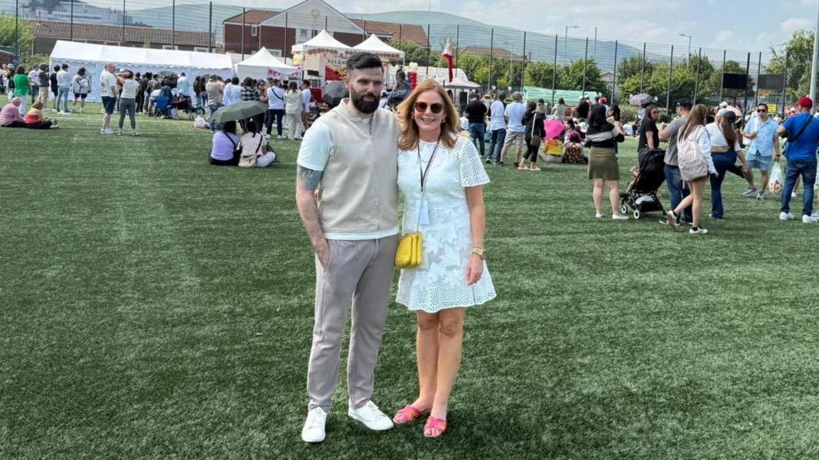 A man and woman stand on a green Astroturf pitch, about a hundred people stand behind them. A large stage can be seen in the background. The man wears a white t-shirt with a cream vest over it with grey trousers and white shoes. He has shirt brown hair and a beard. The woman wears a small white dress, yellow handbag and Pink sandals. She wears brown sunglasses and has long brown hair.