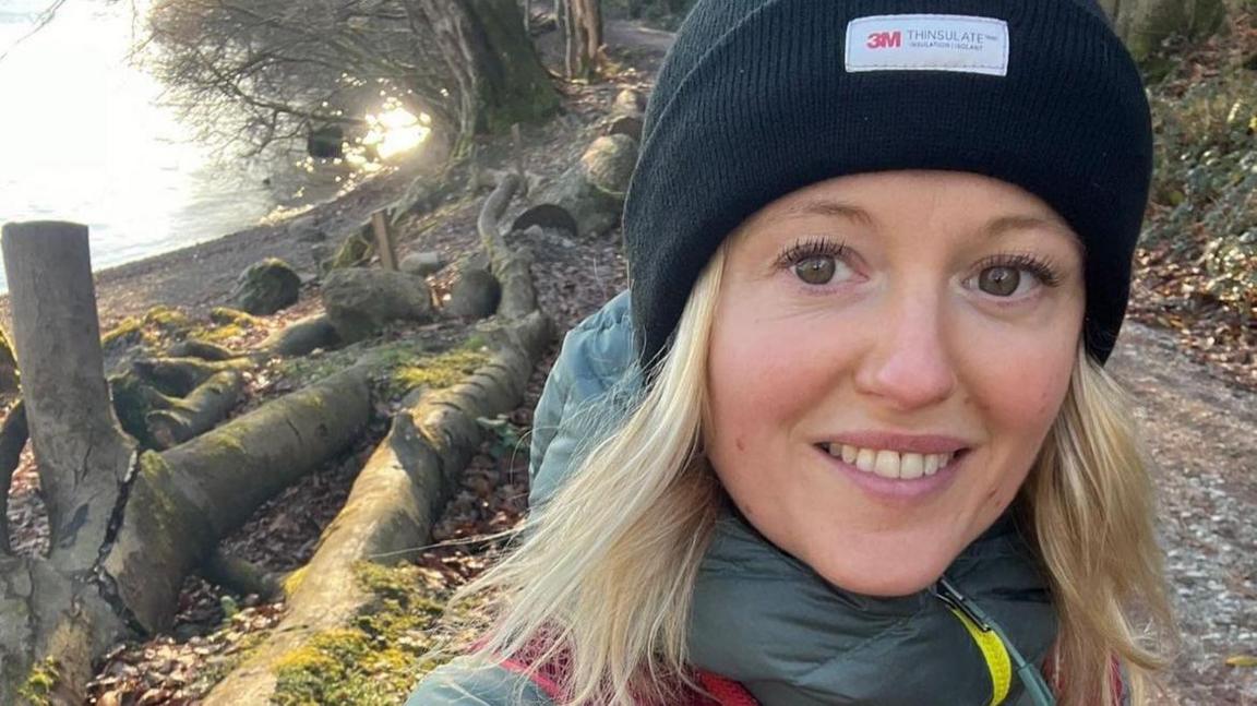 Victoria Bond, a woman with blonde hair and wearing a black woolly hat, smiling at the camera. There are trees and a path in the background.