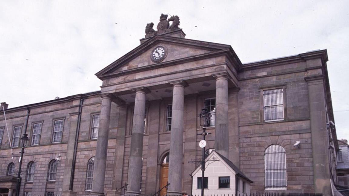 A close-up of Omagh courthouse on a cloudy day. The large, grey stone building has arched Georgian windows and a Palladian portico with a large clock.