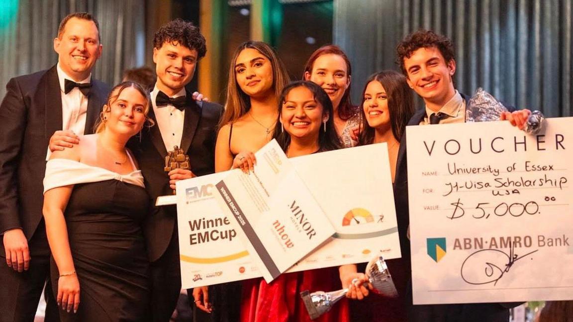 A group of smartly-dressed students smiling with three large cards that indicate their victory and their prize money