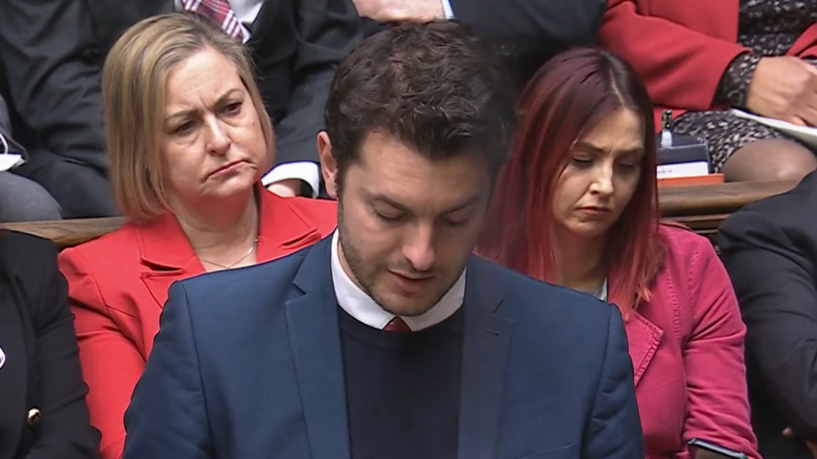 Jack Abbott speaks in the House of Commons during Prime Minister's Questions. He looks down at notes he holds in his hands while he talks. MPs behind him look on. Abbott has short dark hair and wears a navy suit with a black jumper underneath, white shirt and red tie.