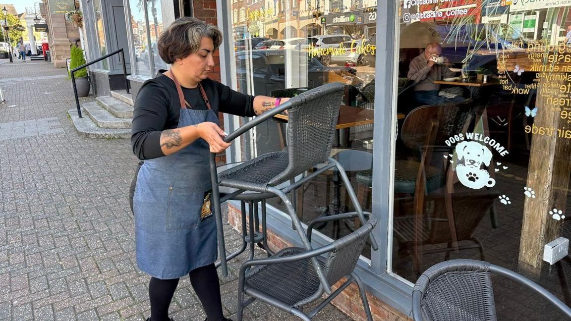 Maria Samaei with medium-length dark hair, wearing a black T-shirt and trousers covered by a blue apron. She is lifting a grey chair which had been stacked on another grey chair, while standing on a grey block-paved pavement next to the glass frontage of a cafe.