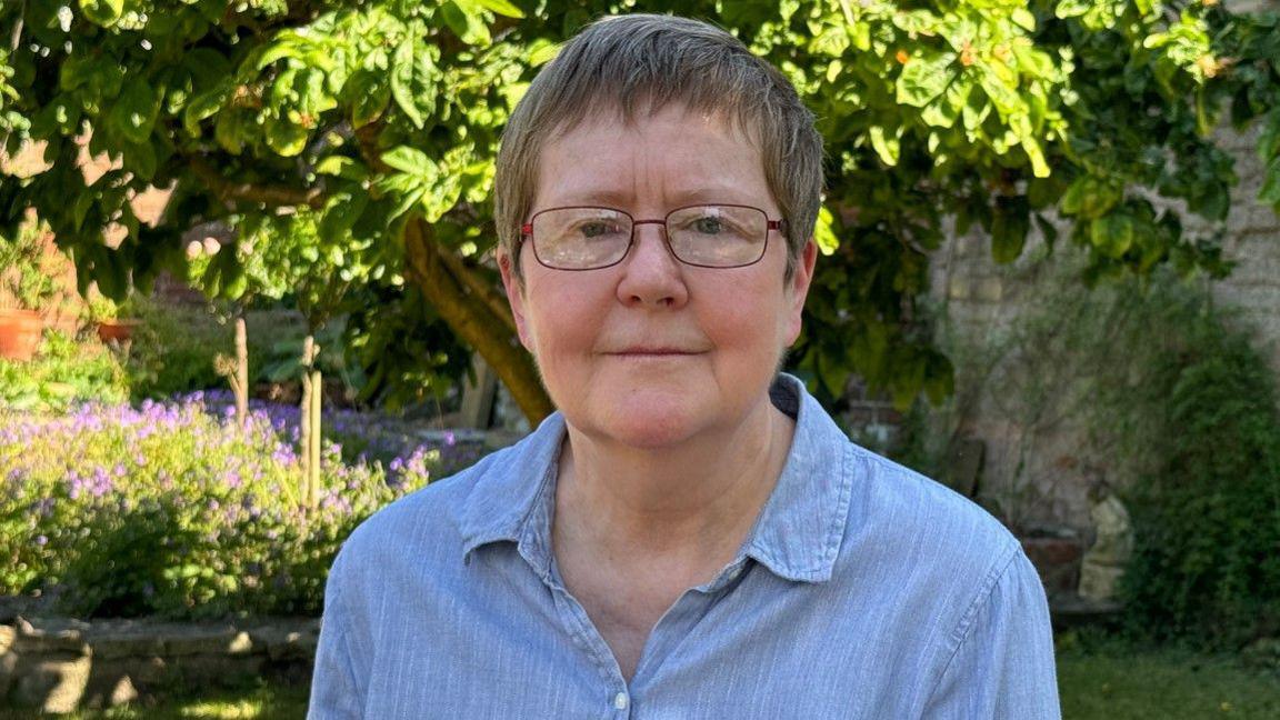 A woman with dark hair and glasses is looking at the camera. She is wearing a blue top. She is standing in a garden with a stone wall, surrounded by trees and flowering plants in the background.