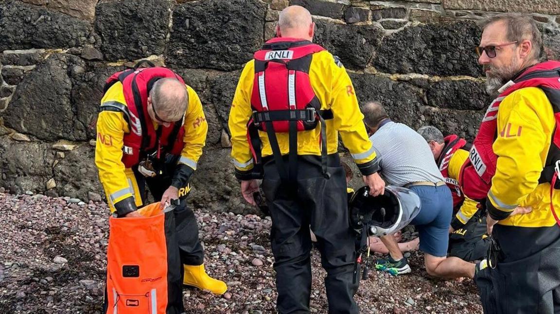 Four RNLI crew members in yellow jackets, black trousers and red life-jackets assessing the scene of an incident off a slip involving a boy, who is sat out of view. A man in blue shorts and a blue shirt is also helping at the incident.