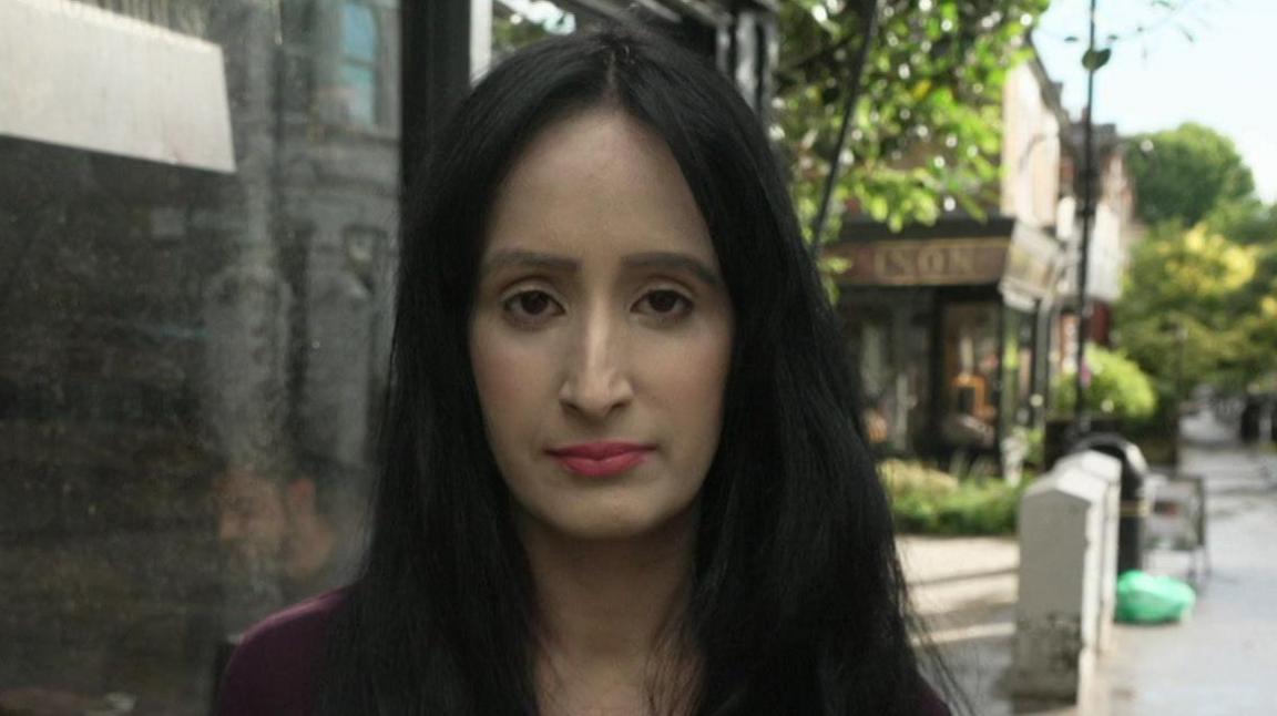 A woman with long dark hair stands on a high street outside a shop. The wet pavement and trees suggest recent rain, and a sign reading “The Axe and Ember” is visible in the background.
