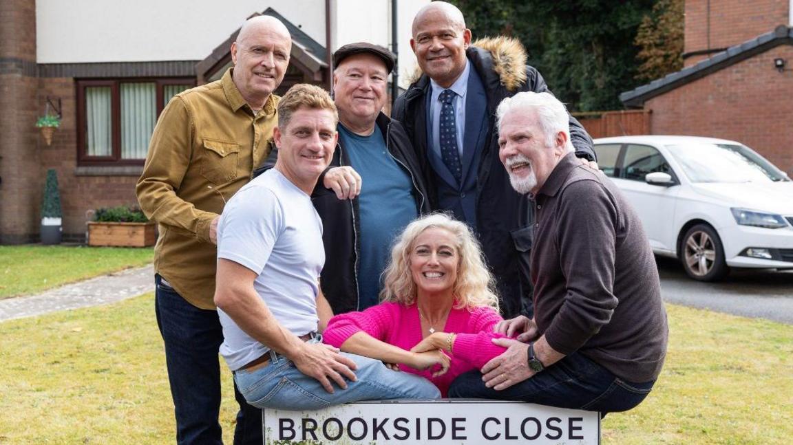 Paul Usher, John McArdle, Michael Starke, Suzanne Collins and Philip Olivier are pictured with the Brookside Close street sign. They are all smiling at the camera. A house with a lawn in front and a white car parked on the driveway are visible in the background