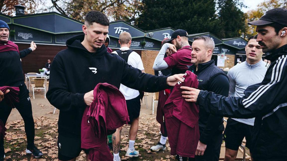 A man in a black hoodie and shorts hands out maroon running t-shirts to people in the park. 