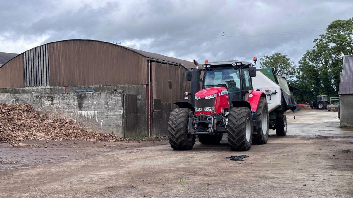 Ryan Taggart driving a tractor with a diet feeder attached. He is driving it in his farm yard which is a concreted yard with sheds. There is a pile of potatoes in the corner which they use as food for the cows. 