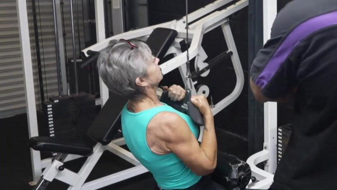 Ms Smith sits on a bench in front of pulldown machine. She pulls down a weighted handle and holds it in front of her chest. A gym instructor coaches her from the side.
