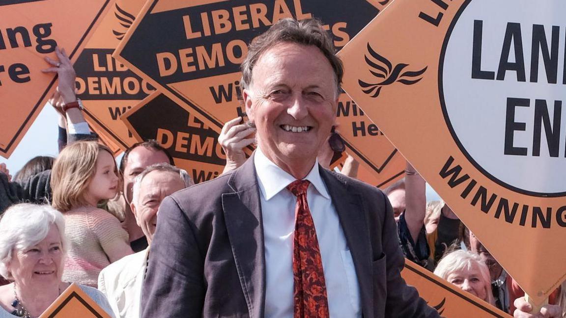 St Ives MP Andrew George smiling with dark brown hair and white sideburns, wearing a grey suit, white shirt and red rose tie with people in the background smiling and hold triangle signs saying LIBERAL DEMOCRATS