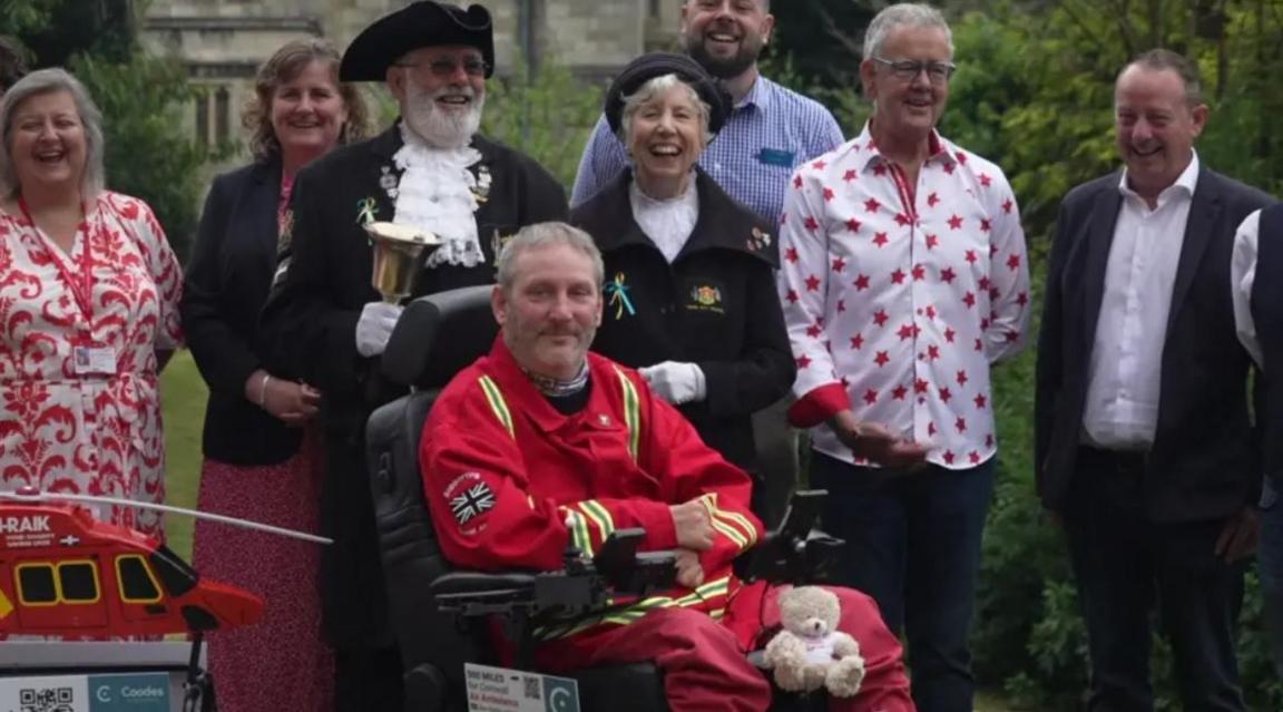 Steven Webb is in the centre of the picture dressed in a red jacket with a Union Jack on the sleeve, around him stand supporters of his cause while a teddy bear perches on his knee and a small Cornwall Air Ambulance replica is to the left. 