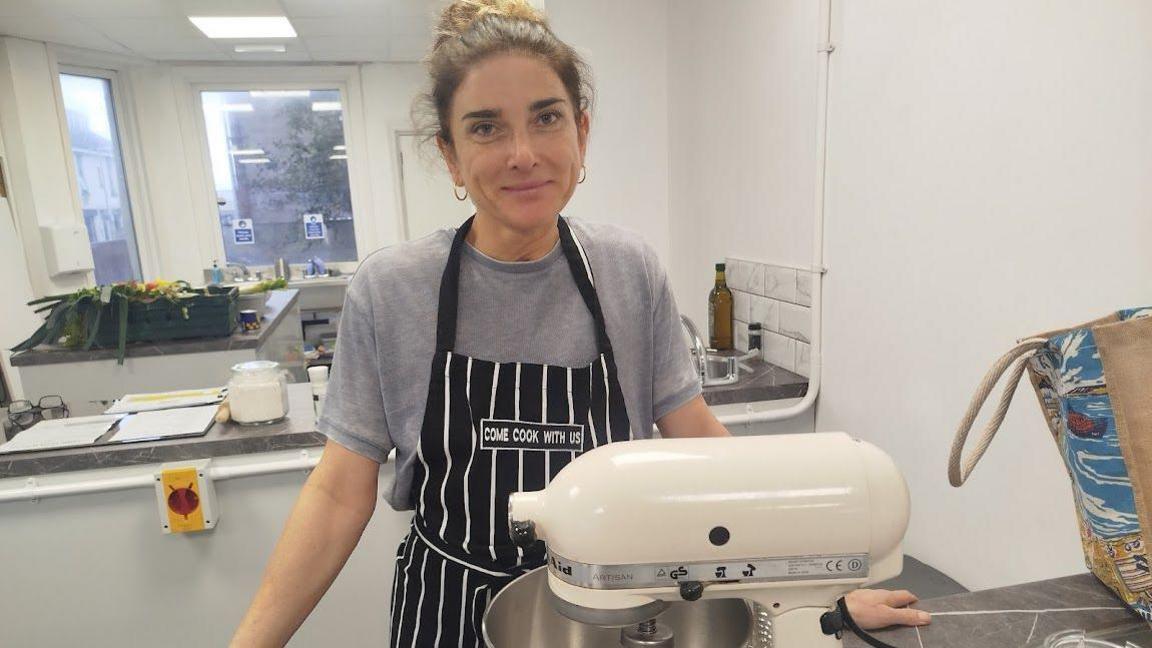 Justine Azzopardi has tied-back black hair and is wearing a black and white striped apron over a grey T-shirt. She stands behind a large mixer and in front of a kitchen worktop.