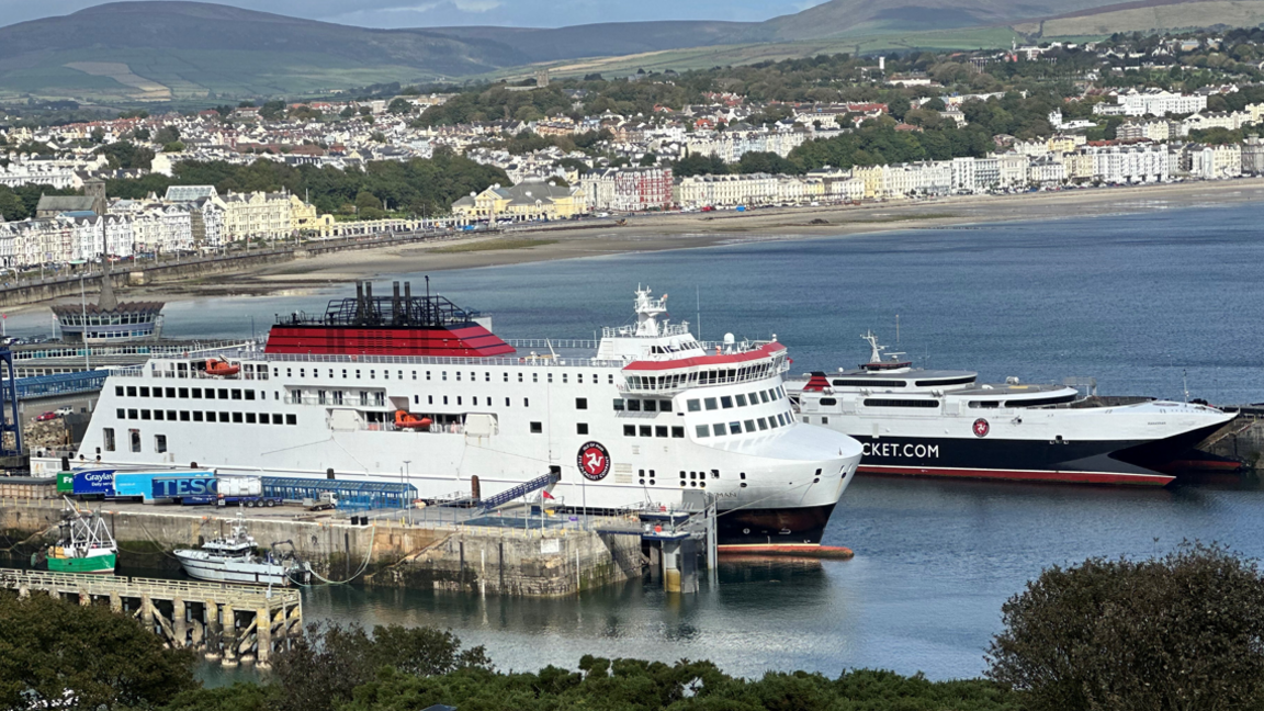 The Manxman and Manannan ferries, which are both painted in the Isle of Man Steam Packet Company's colours of white, red and black, moored in Douglas Harbour. Douglas Bay and the tall white buildings on Douglas Promenade can be seen in the background.