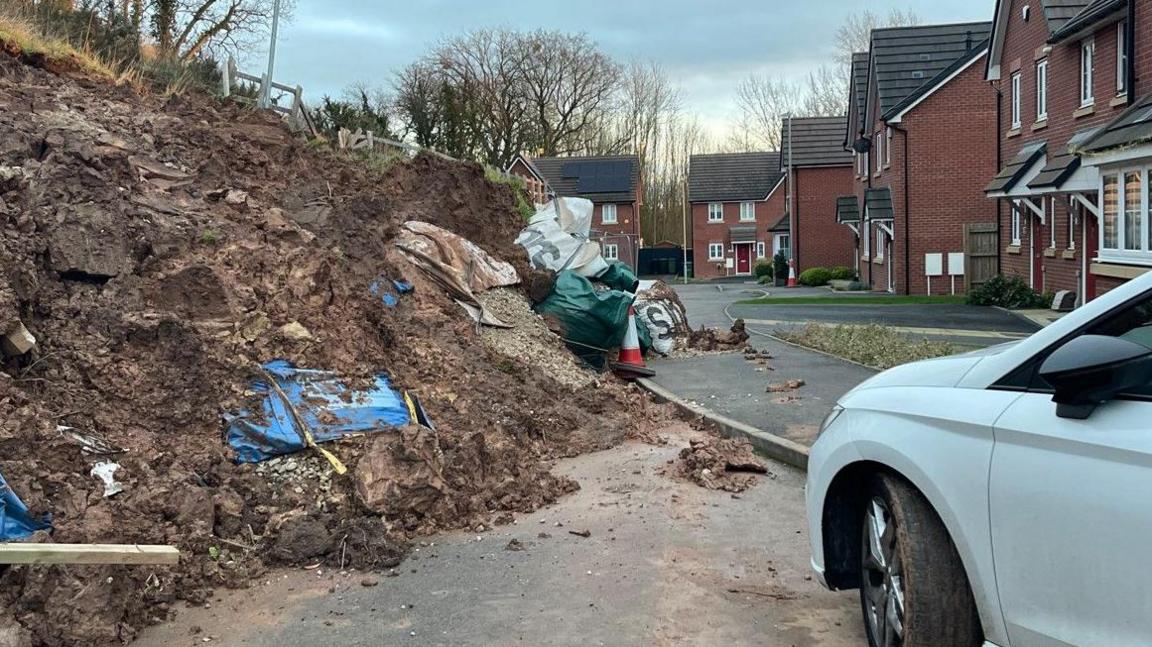 Landslip is visible on the left of the photo. Houses are in the middle and right of the photo in the background. A car is on the right of the photo. 