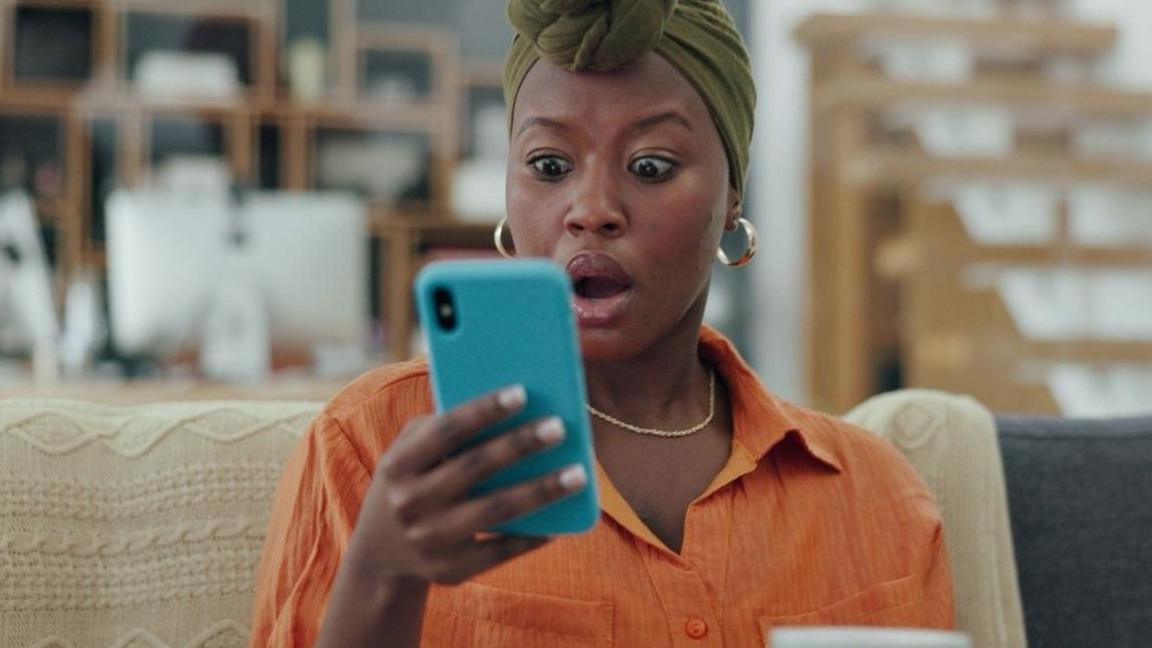 Woman in a green headscarf and orange shirt looks at her phone in shock.