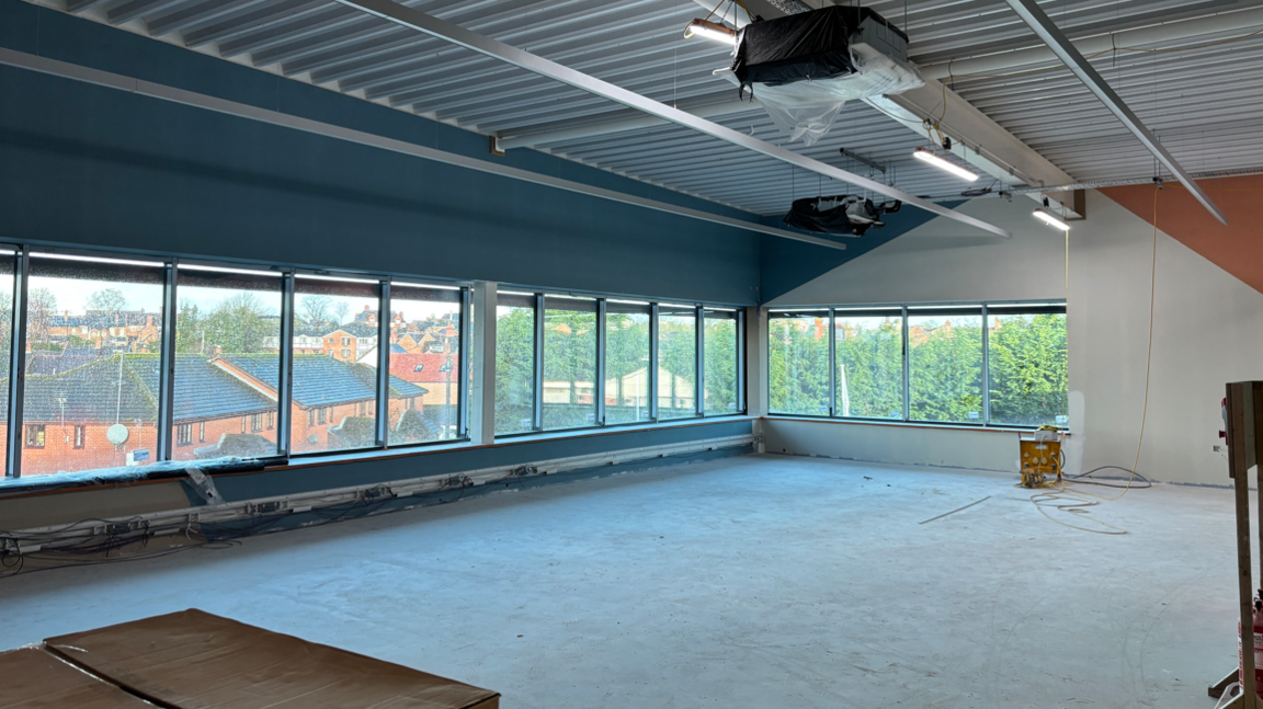 An empty room with a grey floor and windows running along the far wall, with a view of a town beyond. The room is not finished and there are signs of work being carried out on the ceiling and walls.