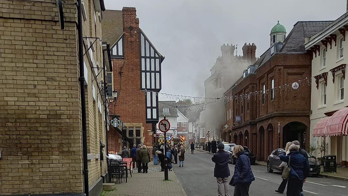 Smoke coming from a building in Saffron Walden, there are a large number of people in nearby streets looking at the fire and you can see fire engines in the distance. You can see a number of buildings and street signs. 