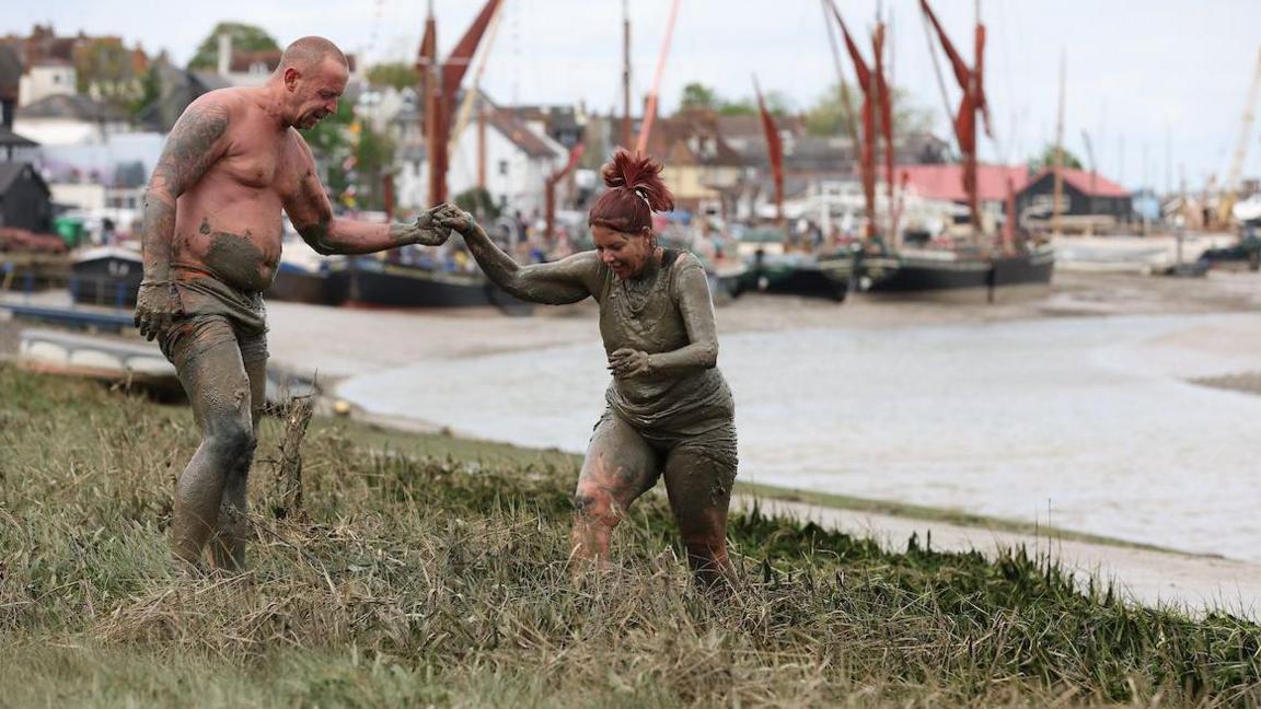 A woman covered in mud being held by the hand by a man who is helping her up a grassy river bank. The water and mud of the estuary is in the background... as are boats on the mud and buildings