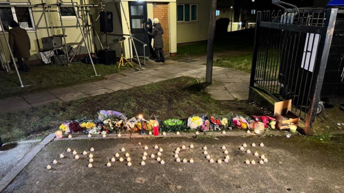 Small candles arranged to spell out the word Marcus on the ground. Floral tributes are behind them on the ground.
