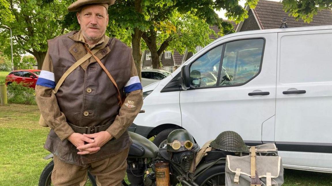 A man in old uniform standing in front of a bike. A white van is behind that and two cars are in the background.