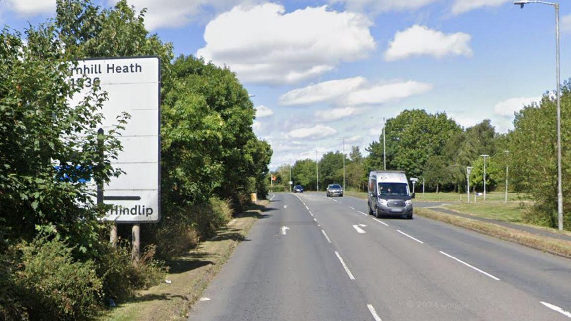 A road with trees down either side and a couple of cars travelling along it and a white road sign on the left hand side