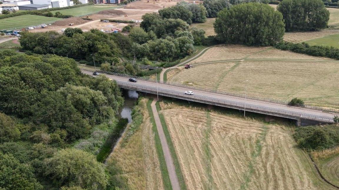An aerial view of a road bridge over a river surrounded by trees and fields.