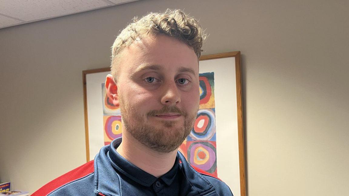 Will pictured looking into the camera and smiling slightly. He's wearing a blue tracksuit top with red shoulders. He's indoors, inside a classroom which has beige walls and a white ceiling. There's a framed piece of modern art behind him. 