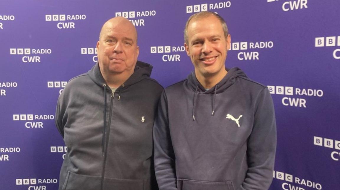 Neil Jeffs on the left is standing next to younger brother Paul, who is smiling at the camera. They are in front of BBC Radio CWR branding.  Neil is bald and wears a light blue hoodie. His brother has short dark to gray hair and smiles to the camera and also wears a light blue hoodie.