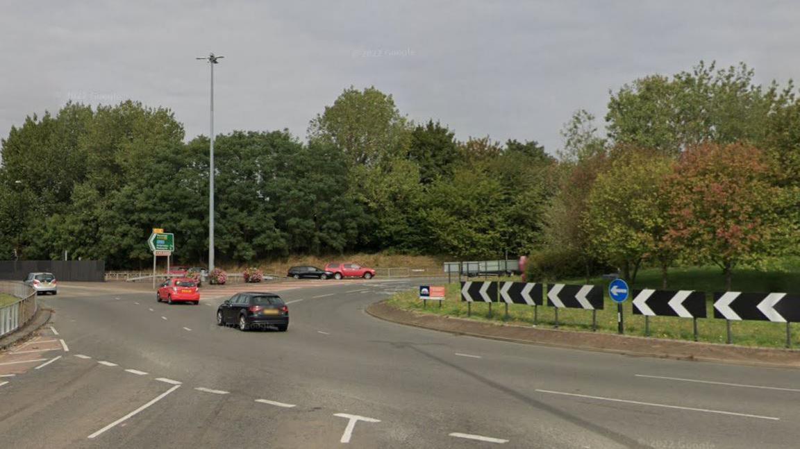 A roundabout connecting two large roads. Black and red cars can be seeing driving on the roundabout and roads.