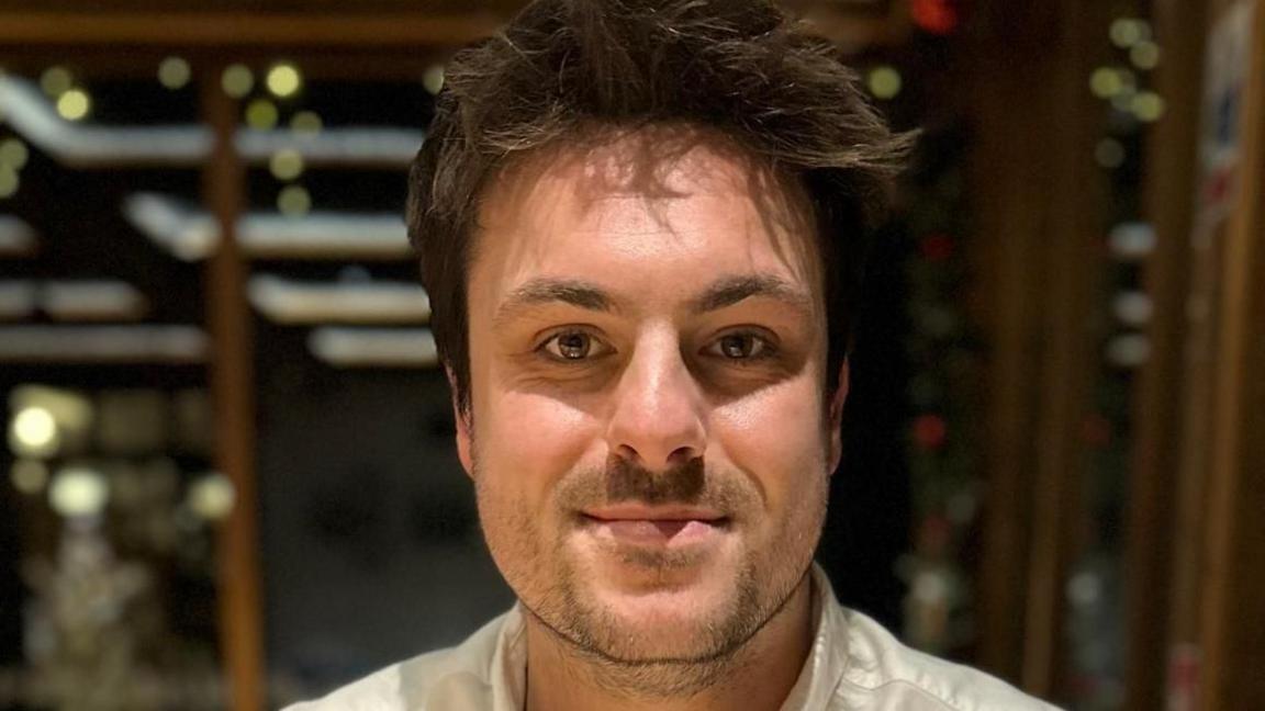 Louis Fields looks directly into the camera in a close up image of his face. He has dark hair and stubble, and is wearing a white shirt.