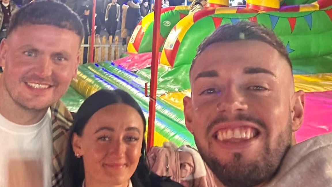 A selfie taken by a man with short brown hair and shaved sides smiling into the camera and wearing a nose ring next to another men with short brown hair and a woman with long black hair. Behind them is a colourful inflatable.
