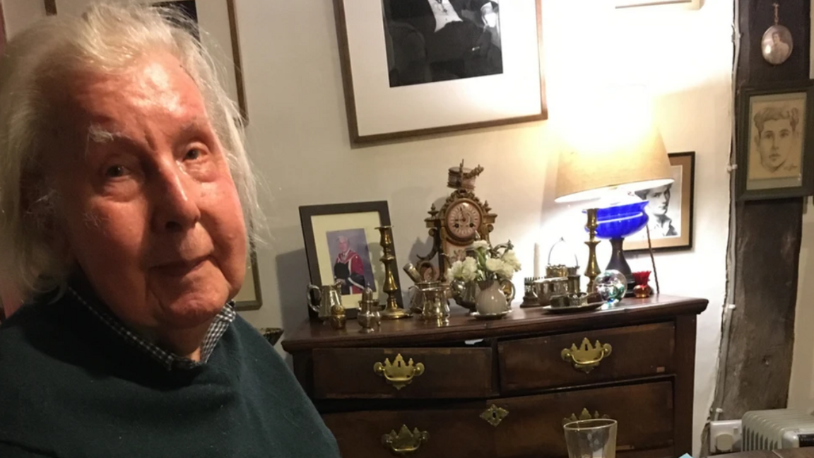 A close-up of Ronald Blythe, who is shown to the left of the image. He is standing in a room in front of a chest of drawers on which there are various items.