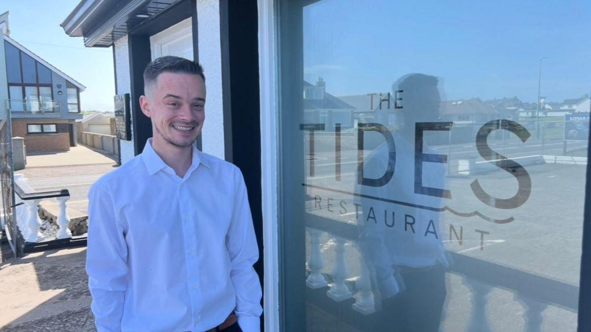 Man wearing a white shirt smiling at the camera. He has dark hair and is standing next to a sign that says Tides