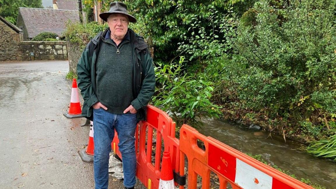 Andy Munro wearing a hat and coat by barriers, near the stream that burst its banks