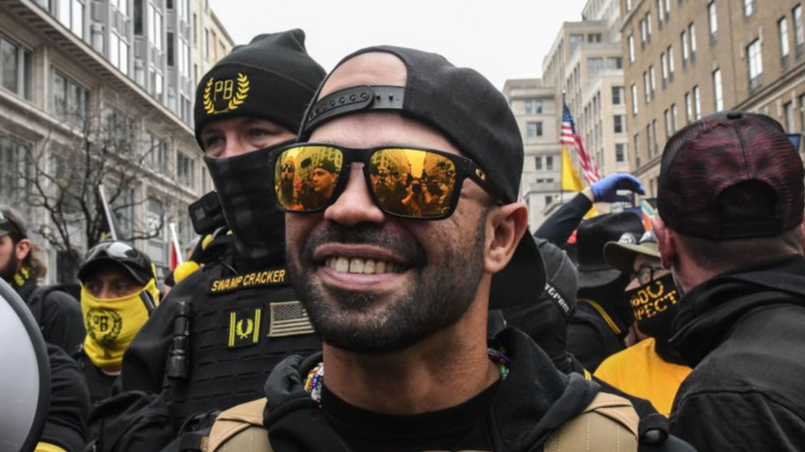 Former Proud Boys leader Enrique Tarrio is shown smiling while wearing mirrored sunglasses and back and yellow protective clothing. He is standing outside Harry's bar in Washington, DC during a protest on 12 December, 2020