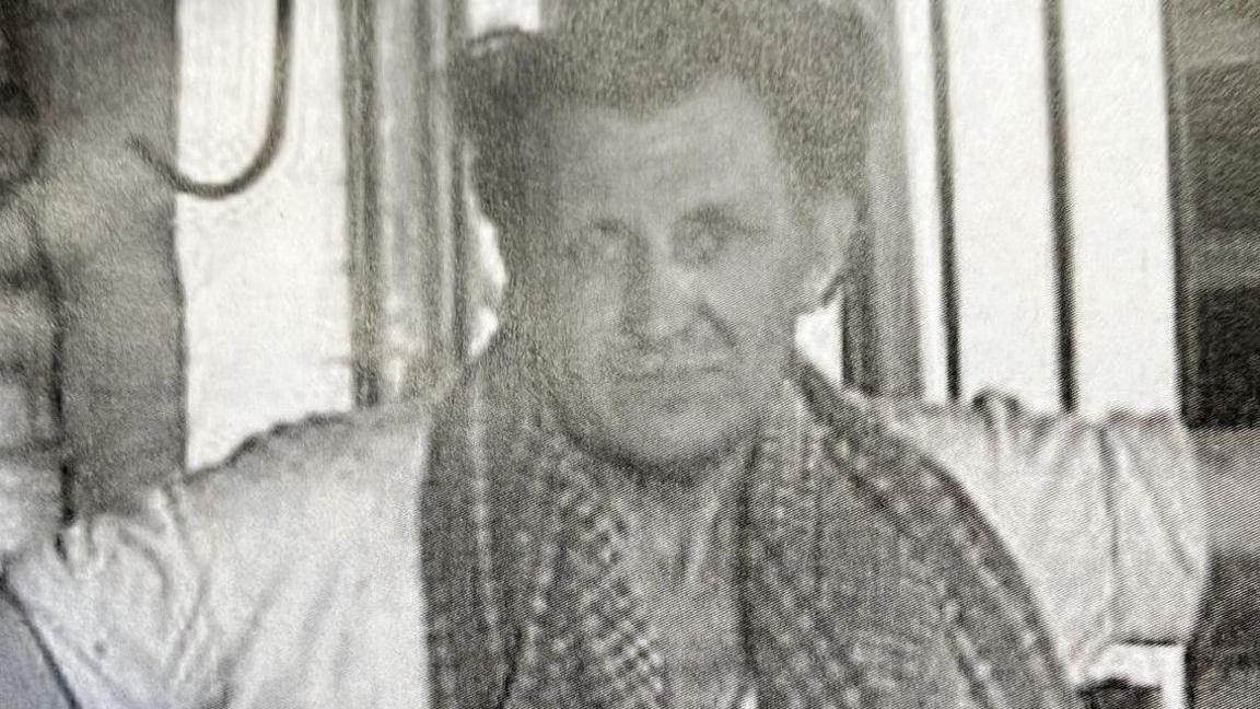 A black-and-white photo of a man who is looking at the camera. He is wearing a white top.