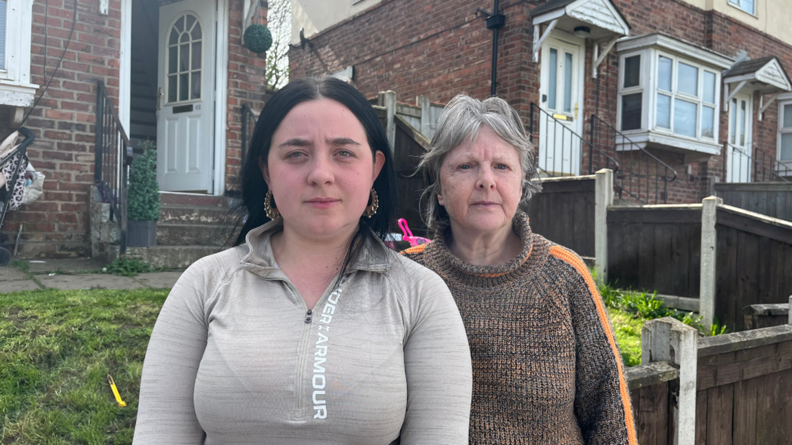 Courtney Mannion and Anne Purchase-Walker standing outside a row of houses. Courtney is wearing a grey top and has black, long hair and has a serious expression. Anne, who has shorter grey hair, stands next to her, wearing a brown jumper.
