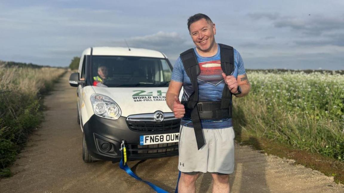 Mike Land in a Superman T-shirt and beige shorts, with a black harness stood in front of a van. He is stood on a dirt track in between two fields. 