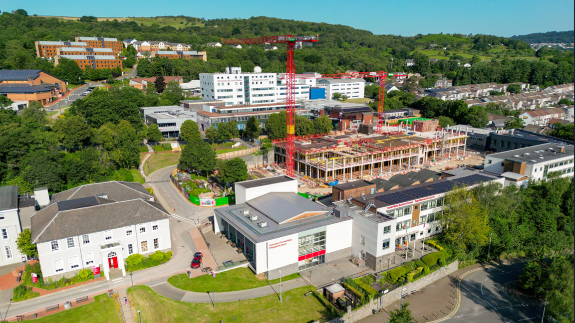 Campws prifysgol yn dangos adeiladau gwyn a gwaith adeiladu a dau graen yng nghanol y safle