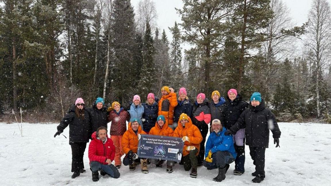 More than a dozen people together in the snow, with some holding a placard saying "we survived the UHCW Charity Arctic Survival Challenge". Trees are behind them.