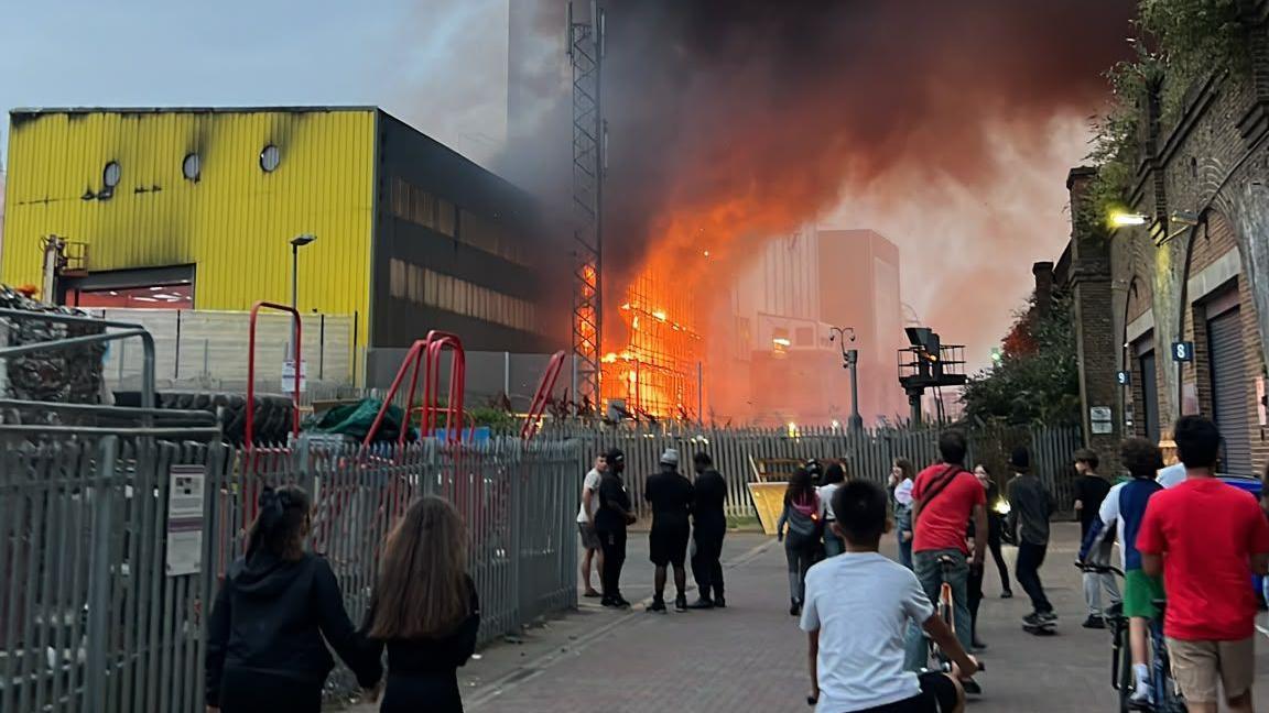 A crowd of bystanders gathers on a street to watch a large blaze sending bright orange flames and thick dark smoke into the sky behind a metal security fence, beside a yellow industrial building and brick railway arches.