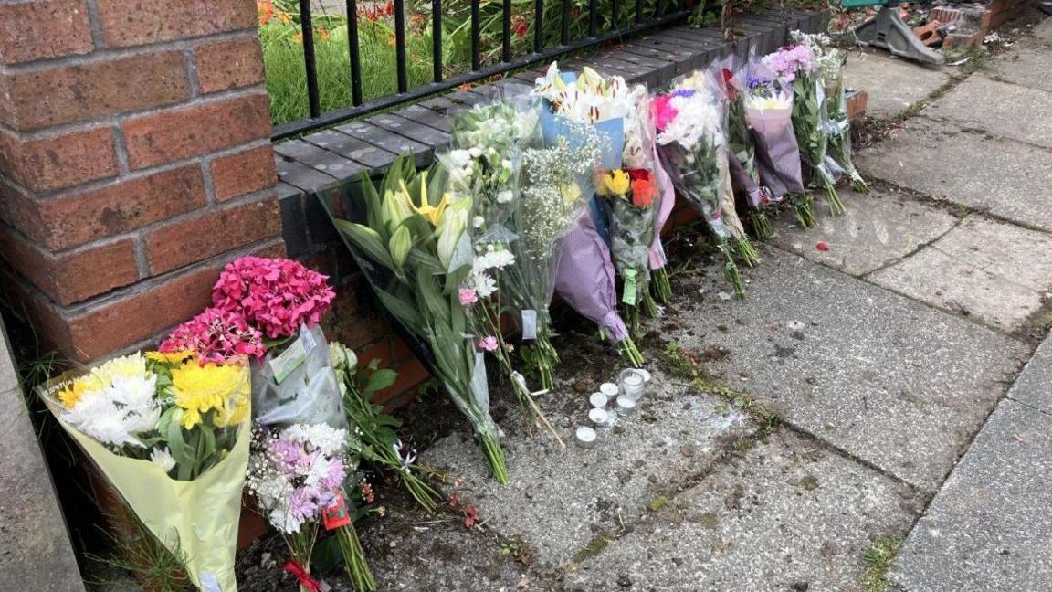 A row of bunches of flowers have been left on the ground near the crash scene. A number of tea lights have been placed in front of the flowers.