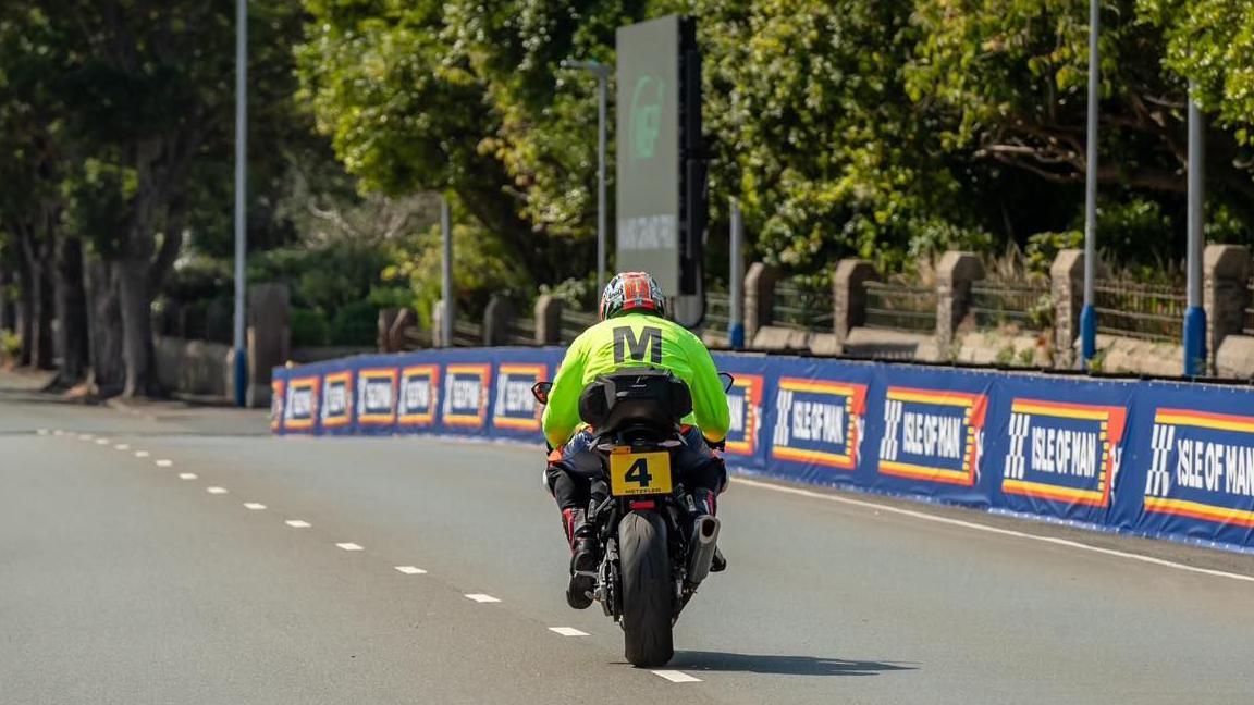 A travelling marshal sets off down Glencrutchery Road. He is wearing a bright yellow jacket with a large M in black lettering on the back. Blue banners with Isle of Man written on them line the right-hand side of the road.