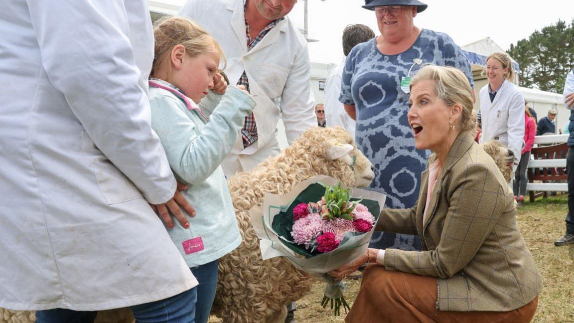 The Duchess of Edinburgh holds a colourful bouquet of flowers while talking to a young girl as she kneels next to a sheep at the Royal Cornwall Sheep.