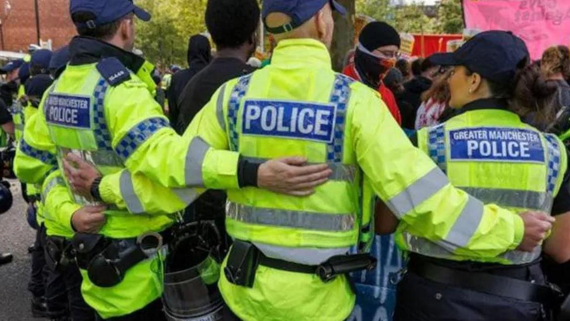 Police officers at a protest stand in a row with their arms around each other to make a barrier. they are wearing hi vis jackets with Greater Manchester Police written on the back and have belts with handcuffs and batons around their waists.