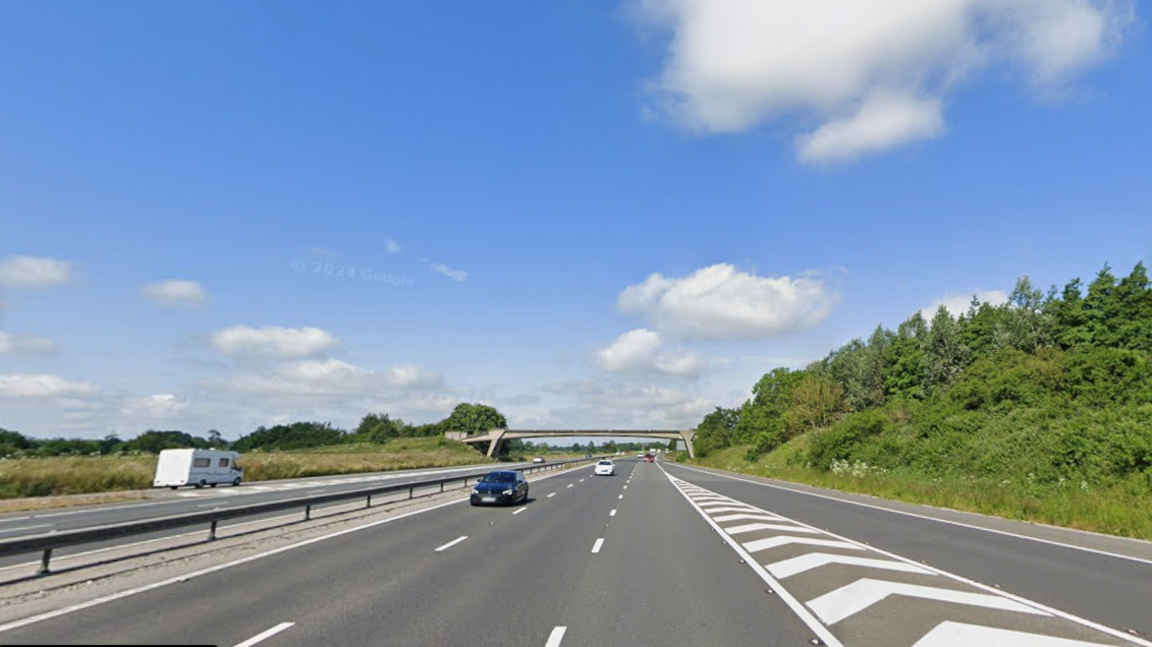 The photo shows a section of motorway as seen from a car. The beginning of a slip lane with black and white arrow markings can be seen to the right and there is a slim concrete bridge passing over the motorway in the background. The sky is blue and dotted with clouds
