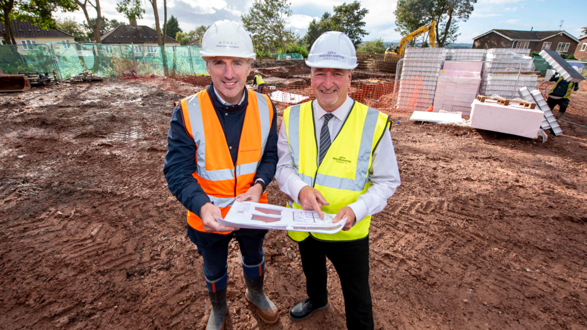 Two men wearing hi-viz tabards and white hard hats, and holding a set of plans, while standing on the soil where the building will be constructed. 