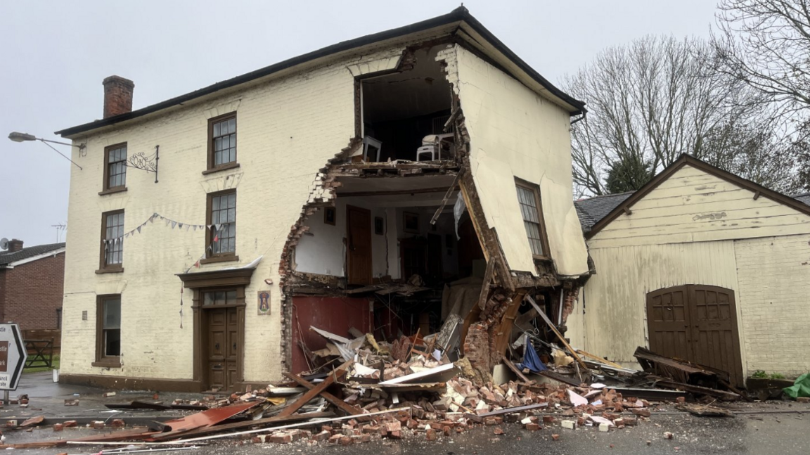 A cream coloured three-storey house has heavily damaged on the right side. Rubble is all over the road and a large hole is now in the house.