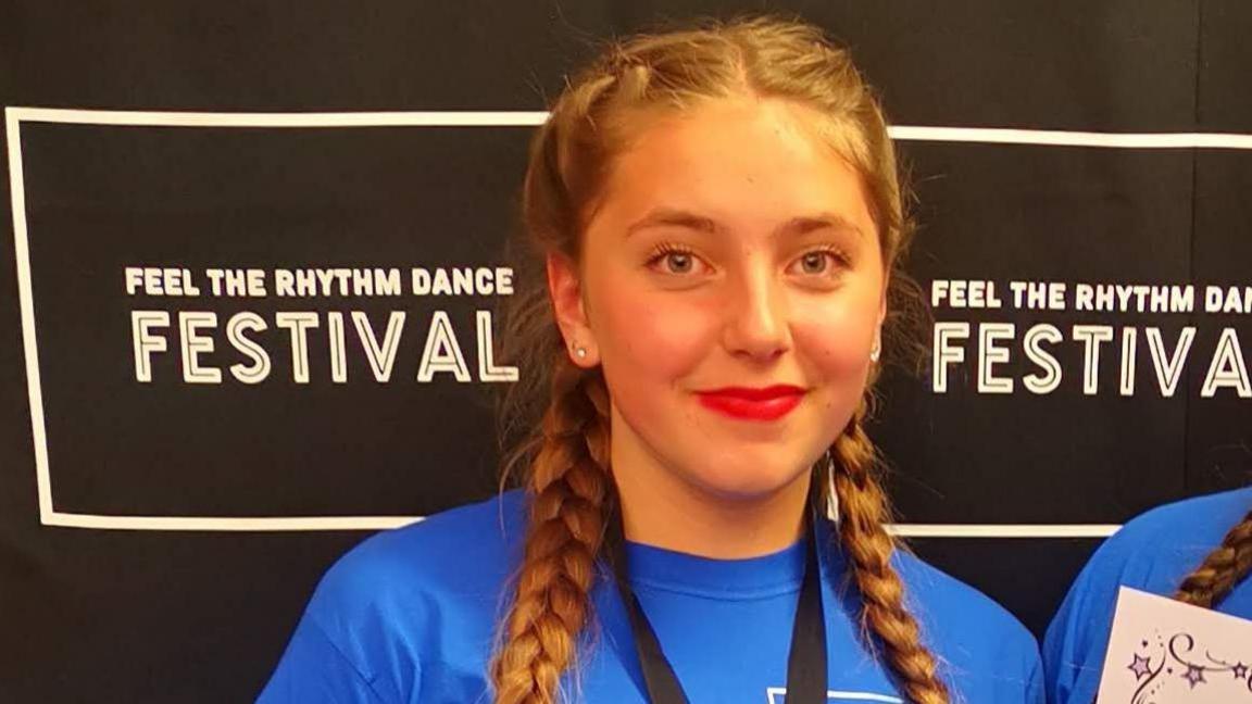 Chloe smiles at the camera posing after coming 3rd in the 'feel the ryhthm dance festival. She has red lipstick on and has her hair in two dutch plaits. She is wearing a blue T-shirt and a black lanyard around her neck.