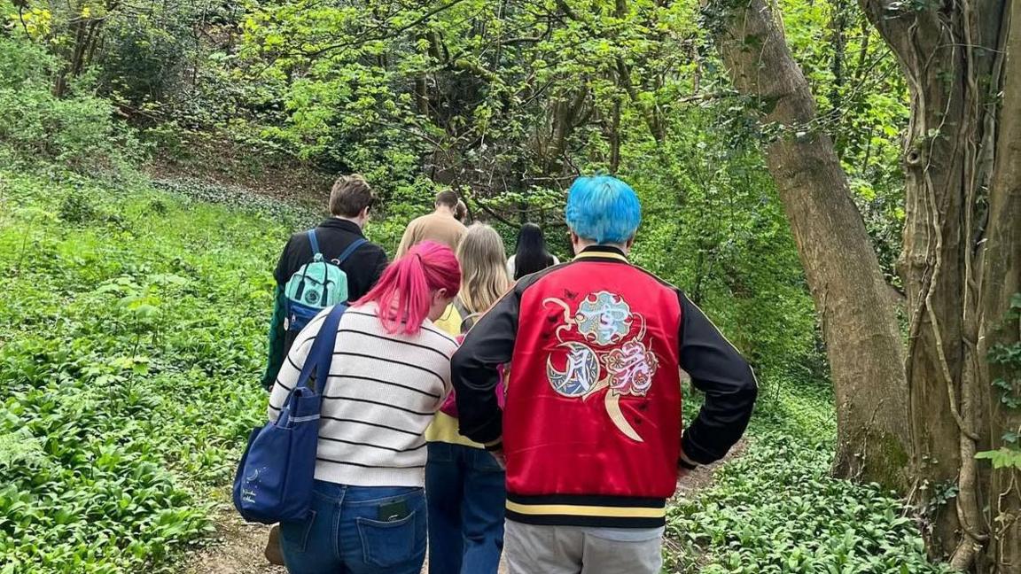 A group of seven people walking down a forest path with the backs to the camera. The two at the back of the group, closest to the camera, have bright coloured hair. There are large trees and wild garlic plants on the forest floor. 